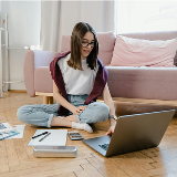 Young woman seated on the floor of an apartment, leaning on a sofa, while working on a laptop placed next to her on the floor.