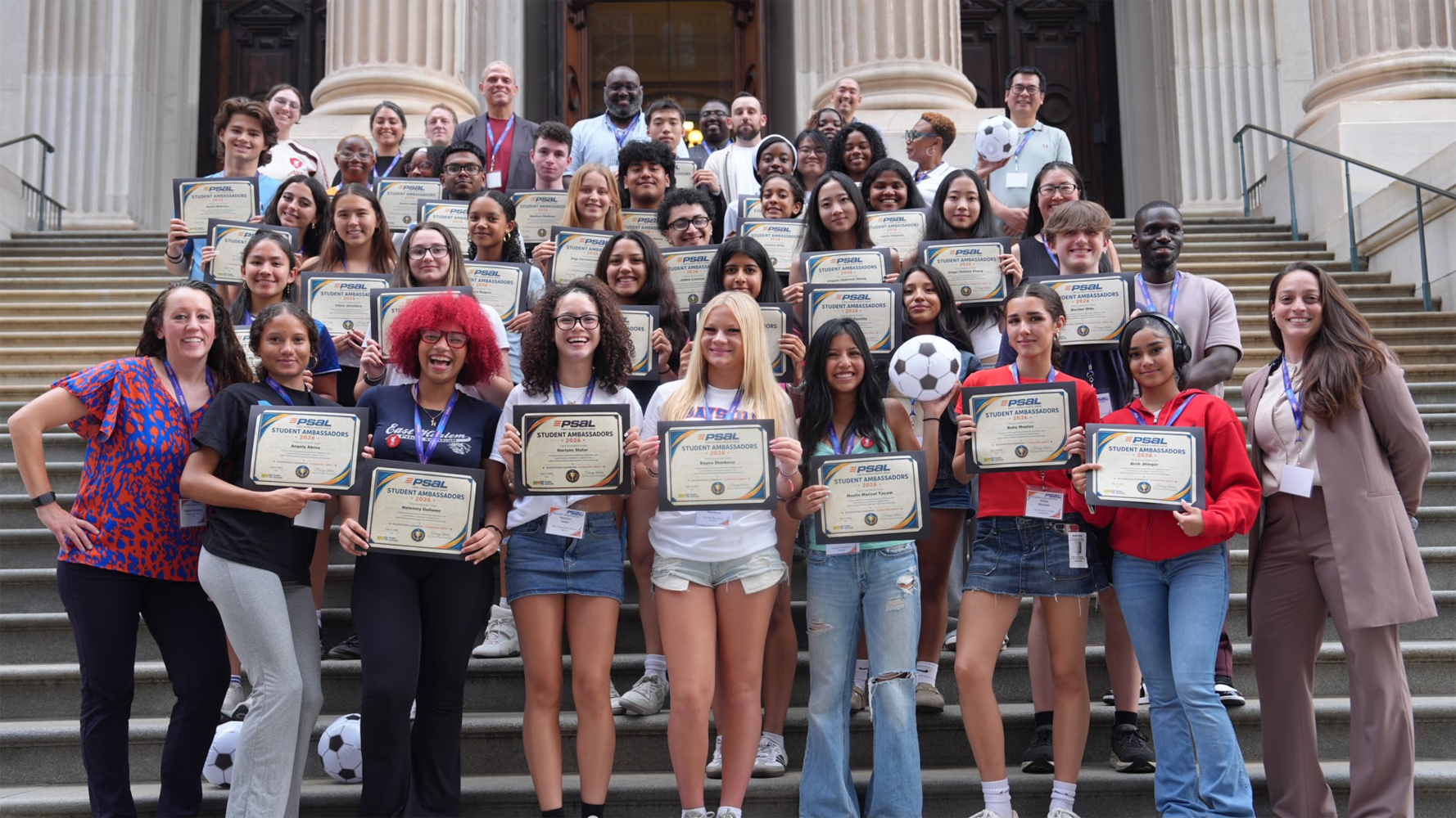 Group of high school students standing on a staircase and holding up certificates that read, 'PSAL Student Ambassadors'