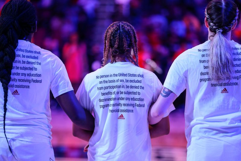 Women's Basketball Players Before Tipoff The backs of three women's basketball players standing arm in arm before tipoff wearing t-shirts containing text from the Title IX legislation
