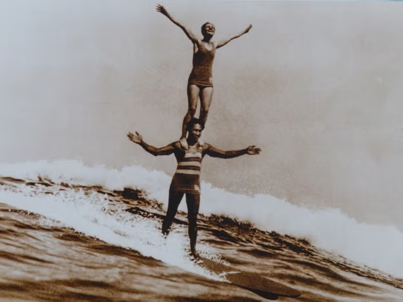 A man, Duke Kahanamoku, surfing with his arms out on a wave, with a woman, Viola Hartman, standing on his shoulders.