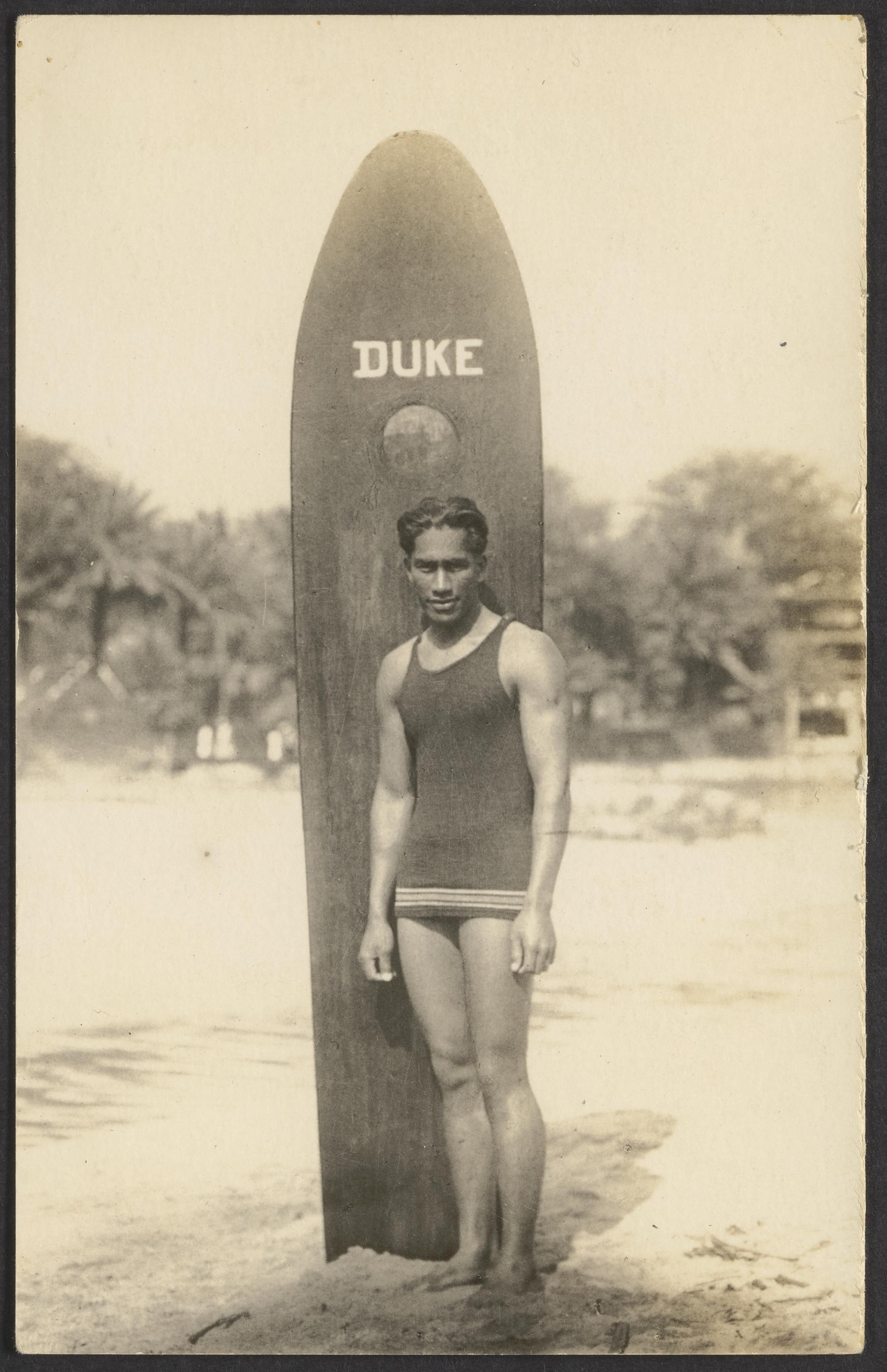 Duke Kahanamoku standing with a wooden surfboard that is planted in the sand and has his name written at the top.