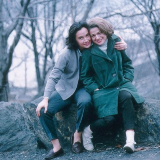 Thea Spyer (left) sitting with her arm around Edie Windsor (right) outside on some rocks with bare trees in the background.