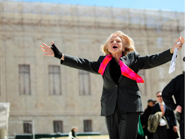 Edie Windsor on the steps of the Supreme Court. She is wearing a dark suit with a pink scarf and holding her arms out to the side in celebration.