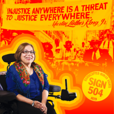 Photo of Judy Heumann in front of a sign reading 'Injustice anywhere is a threat to justice everywhere.'