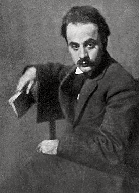 Black and white photograph of Kahlil Gibran, a man with dark hair and a moustache wearing a black coat, sitting down with a book in his hand.