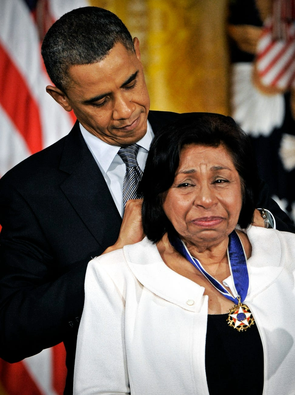President Barack Obama puts the Presidential Medal of Freedom, a star-shaped medal with a blue ribbon, around the neck of Sylvia Mendez. Mendez is wearing a white jacket and black top, and looks like she is tearing up.