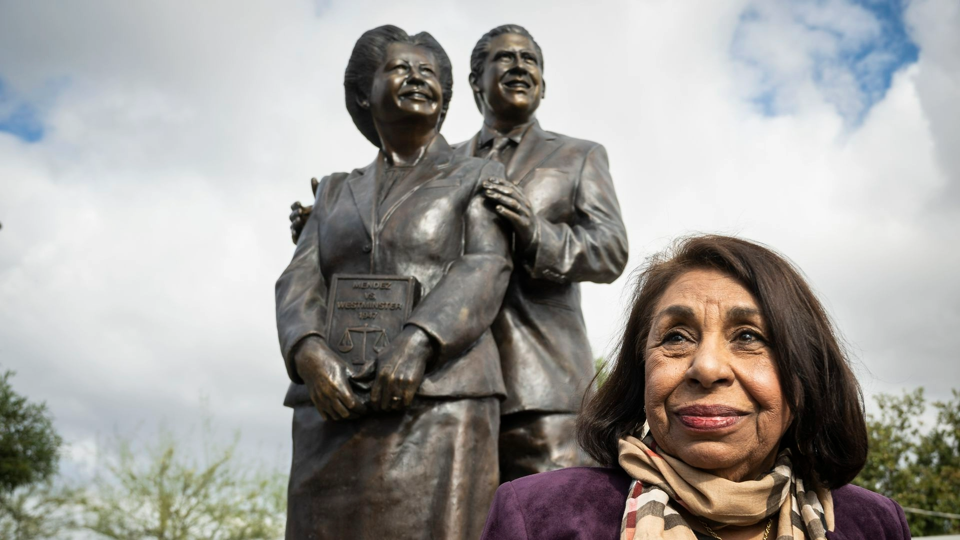 Color photograph of Sylvia Mendez as an adult standing in front of a statue of her parents Gonzalo and Felicitas. Sylvia is smiling, and there are clouds and trees in the background.