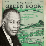 Black and white photograph of Victor Hugo Green in front of a copy of the Fall 1956 copy of "The Negro Travelers' Green Book" which features a green-toned illustration of two people driving in a car.