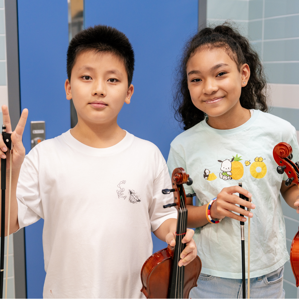 Pair of middle school-aged students holding violins and smiling at the camera