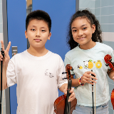 Pair of middle school-aged students holding violins and smiling at the camera