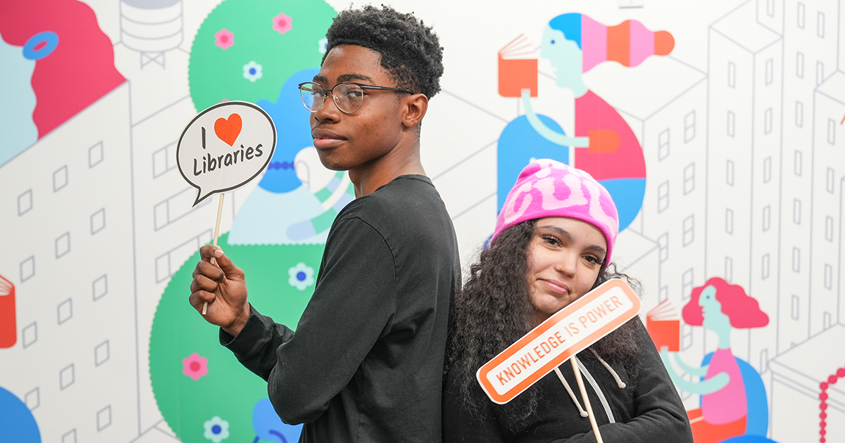A teen boy (left) and girl (right) pose together in front of a colorful background featuring an illustrated figure reading a book, holding up signs that read 'I Love Libraries,' and 'Knowledge is Power,' respectively.