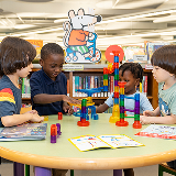 Four students sitting at a table in a library working on a STEAM activity with a bookshelf in the background.