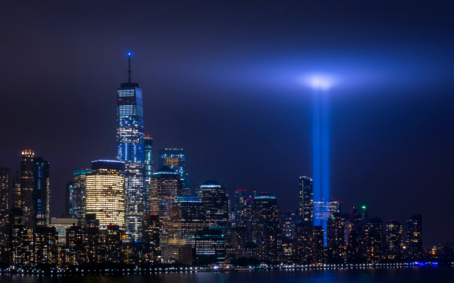 Photograph at night of the NYC skyline with One World Trade Center to the left and two beams of light, memorials to the Twin Towers, glowing to the right.