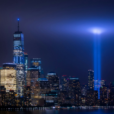 Photograph at night of the NYC skyline with One World Trade Center to the left and two beams of light, memorials to the Twin Towers, glowing to the right.
