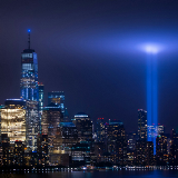 Photograph at night of the NYC skyline with One World Trade Center to the left and two beams of light, memorials to the Twin Towers, glowing to the right.