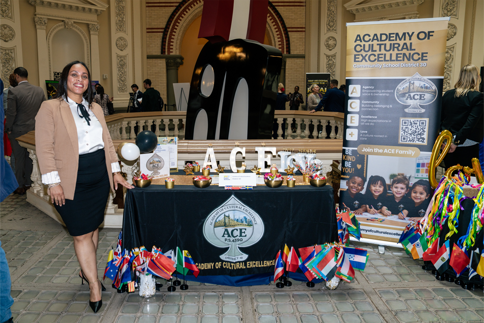 A woman standing on the left of a table display for the Academy of Cultural Excellence featuring dozens of small flags of countries from around the world, and the letters A-C-E in illuminated block lettering.