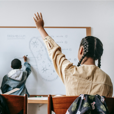 Back of classroom with young girl raising hand while looking on another student writing on a whiteboard as a older woman looks on in approval.