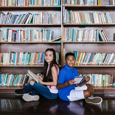 A middle school-aged boy and girl sitting on the floor of a library with books open in their hands.