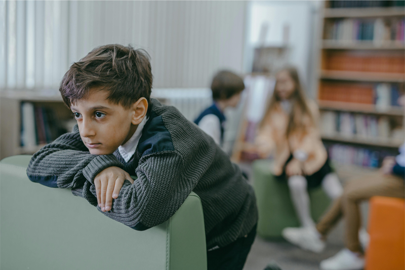 Close-up view of a young adolescent boy, leaning over a chair, looking pensive and adrift, while three other children are conversing amongst themselves in the background, completely oblivious to the boy.