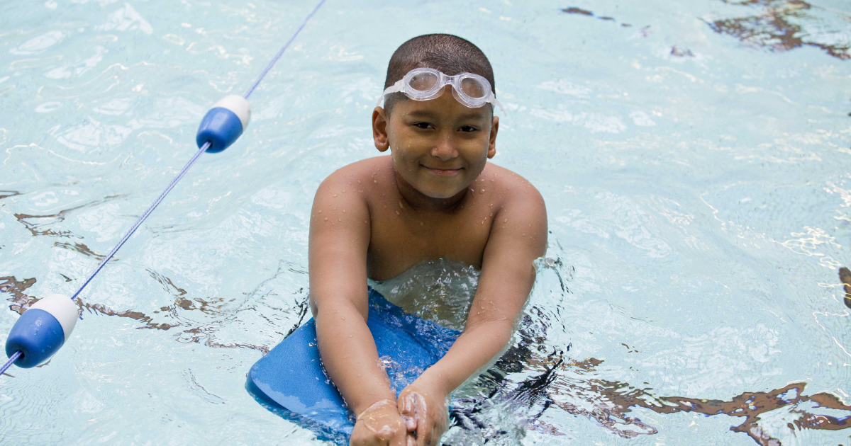 Boy with goggle on his forehead in a swimming pool holding onto a blue board.