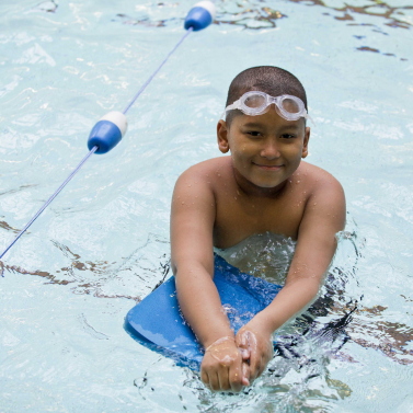 Boy with goggle on his forehead in a swimming pool holding onto a blue board.