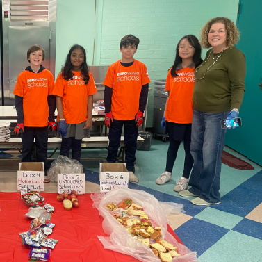 Students in orange shirts and a teacher lined up in a cafeteria behind food waste from their school's lunch sorted into boxes.