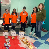 Students in orange shirts and a teacher lined up in a cafeteria behind food waste from their school's lunch sorted into boxes.