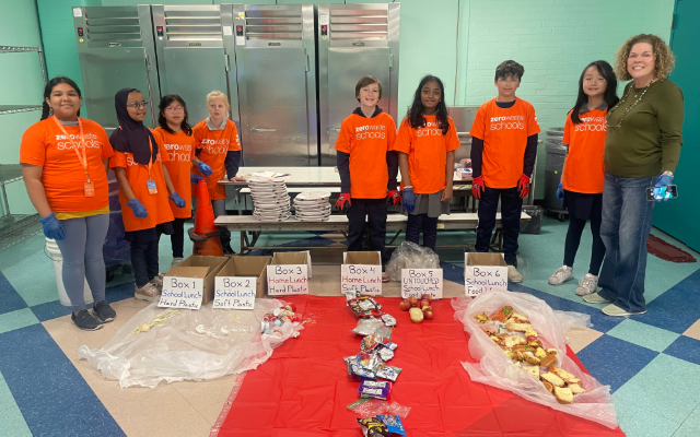 Students in orange shirts and a teacher lined up in a cafeteria behind food waste from their school's lunch sorted into boxes.