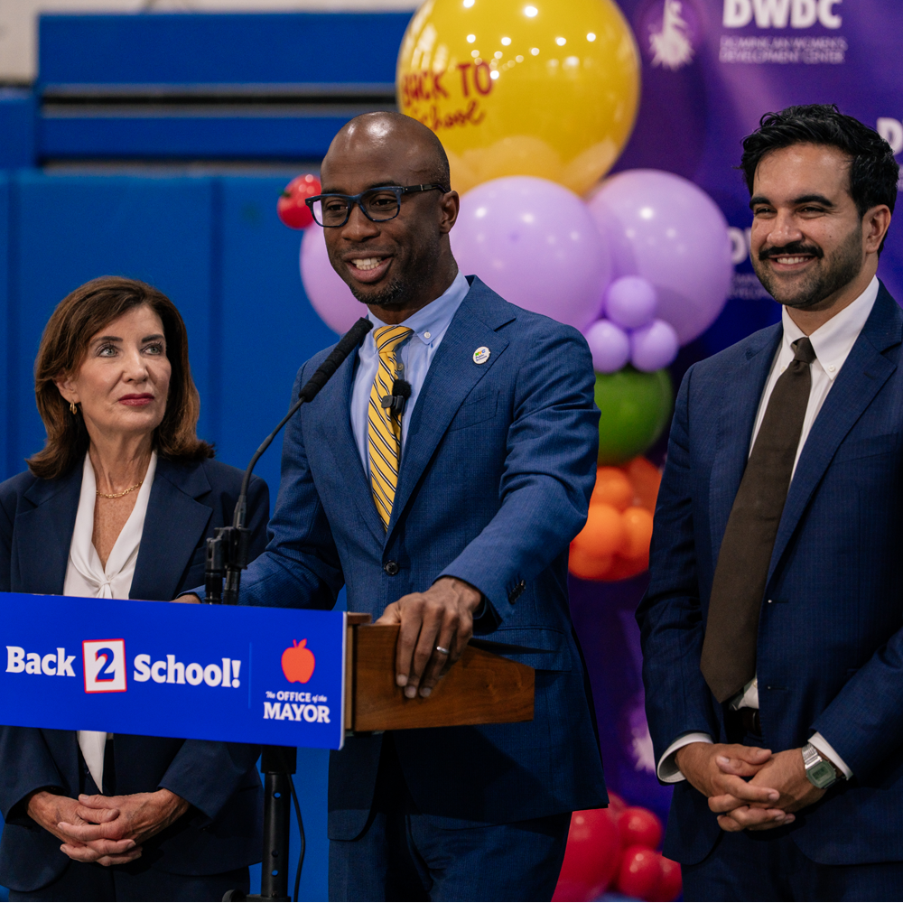 Photo featuring Governor Kathy Hochul (left), Chancellor Kamar Samuels (center), and Mayor Zohran Mamdani (right) standing at a podium and smiling.