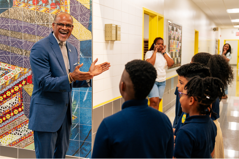 Chancellor David Banks (left) clapping his hands while talking to four young students standing in a school hallway.