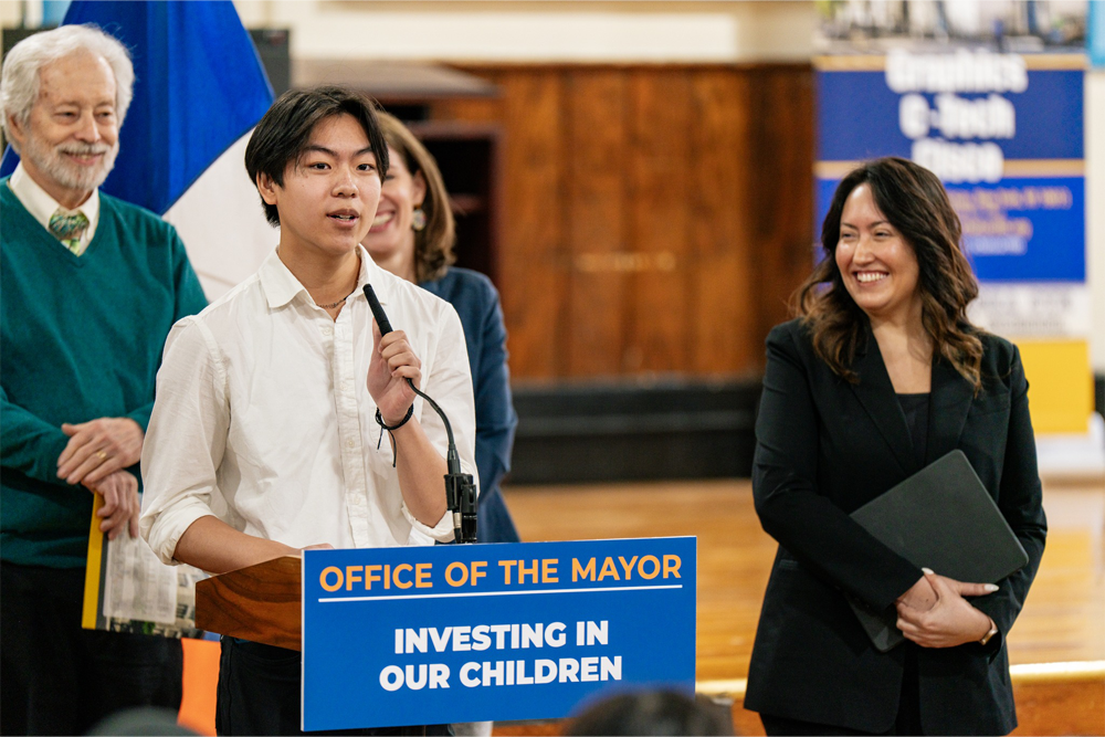 Chancellor Ramos (right) smiling while looking on at a student giving a speech at a podium (left) during a Mayoral press conference.