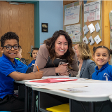 Chancellor Melissa Aviles-Ramos (center) leaning over a table and smiling in between two adolescent students who are also smiling.