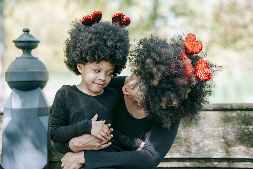 A mother holding her daughter in her arms while sitting on a wooden bench outdoors and wearing Valentine's Day-themed headbands.