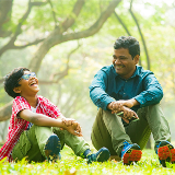 Elementary/middle school-aged child sitting on grass in a park next to an older man, and they are both laughing