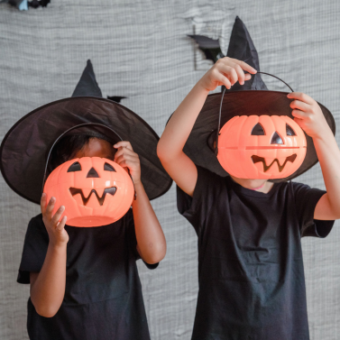 Two children wearing witch hats cover their faces with Halloween pumpkin pails