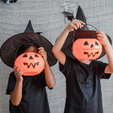 Two children wearing witch hats cover their faces with Halloween pumpkin pails