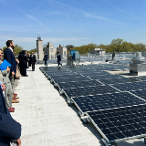 People standing on a roof covered with solar panels with a blue sky and trees in the background.