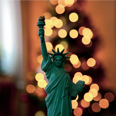 Close-up photo of a miniature figure of the Statue of Liberty; holiday lights can be seen in the background behind Lady Liberty.
