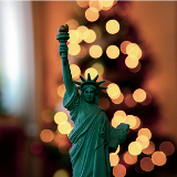 Close-up photo of a miniature figure of the Statue of Liberty; holiday lights can be seen in the background behind Lady Liberty.