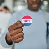 Square cut of a closeup of a person's hands holding up a "Vote" sticker