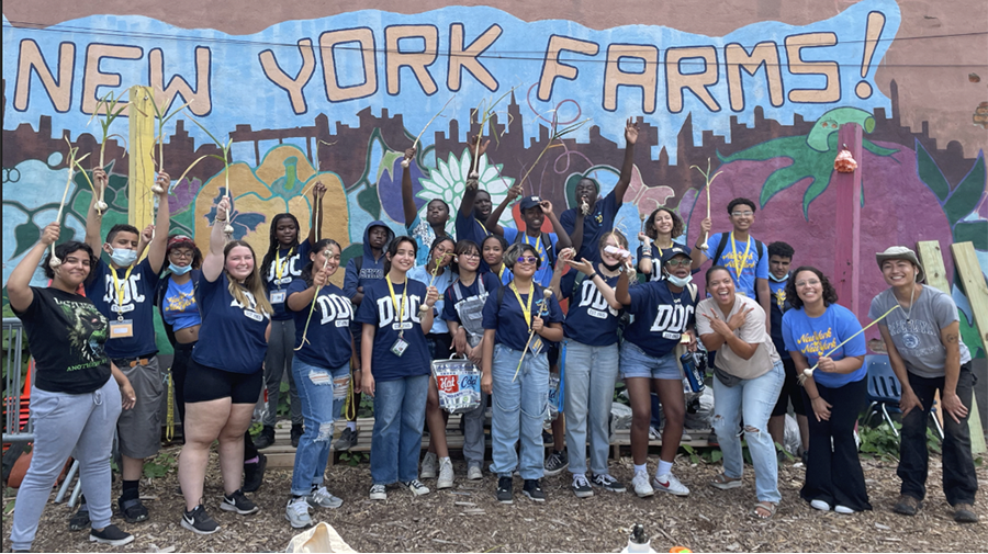 Group of about 25 people posing together for a photo in front of a mural with the phrase, 'New York Farms!' written on it.