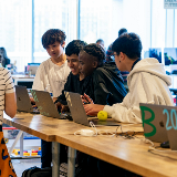 Four students gathered around a table with laptops working together on a computer.