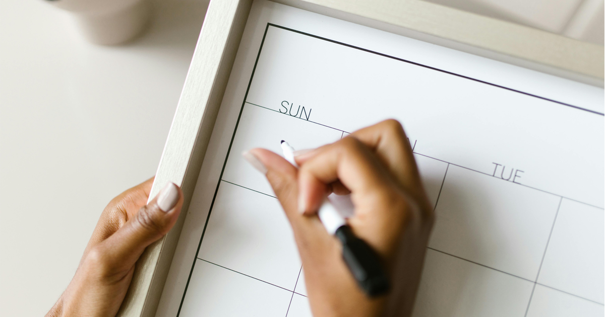 Close-up of a pair of hands starting to write on a dry-erase calendar with a dry-erase marker.