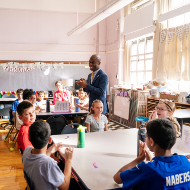 Chancellor Samuels stands in a classroom with smiling students seated at tables with a bulletin board in the background that says 'Reading.'