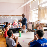 Chancellor Samuels stands in a classroom with smiling students seated at tables with a bulletin board in the background that says 'Reading.'