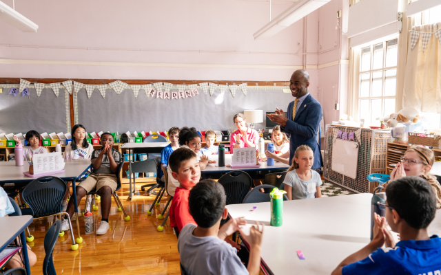 Chancellor Samuels stands in a classroom with smiling students seated at tables with a bulletin board in the background that says 'Reading.'