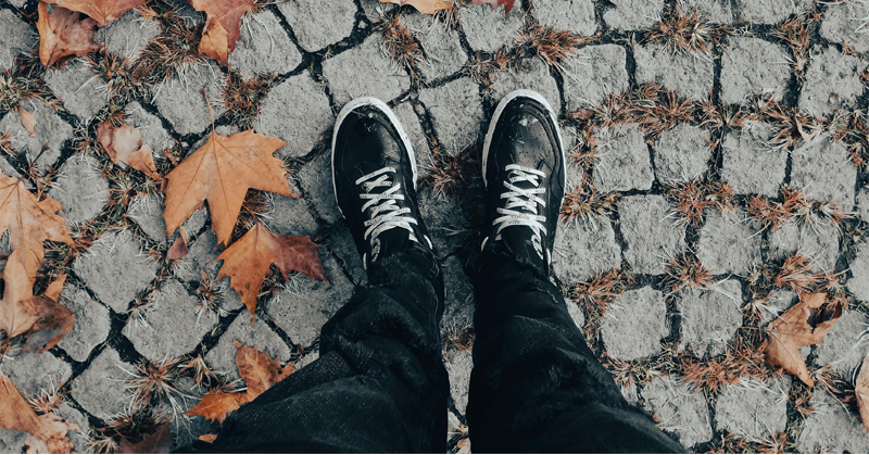 Close-up of a pair of feet (wearing sneakers) that are standing on cobblestone lined with brown dried leaves.