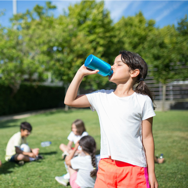 Student with long hair drinking from a reusable bottle in the foreground, while four other middle school-aged children are sitting down in grass facing one another in the background.