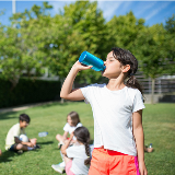 Student with long hair drinking from a reusable bottle in the foreground, while four other middle school-aged children are sitting down in grass facing one another in the background.