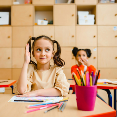 Female student raising her hand while sitting at a classroom desk, and one other students behind her is paying attention to another person or object off-camera.